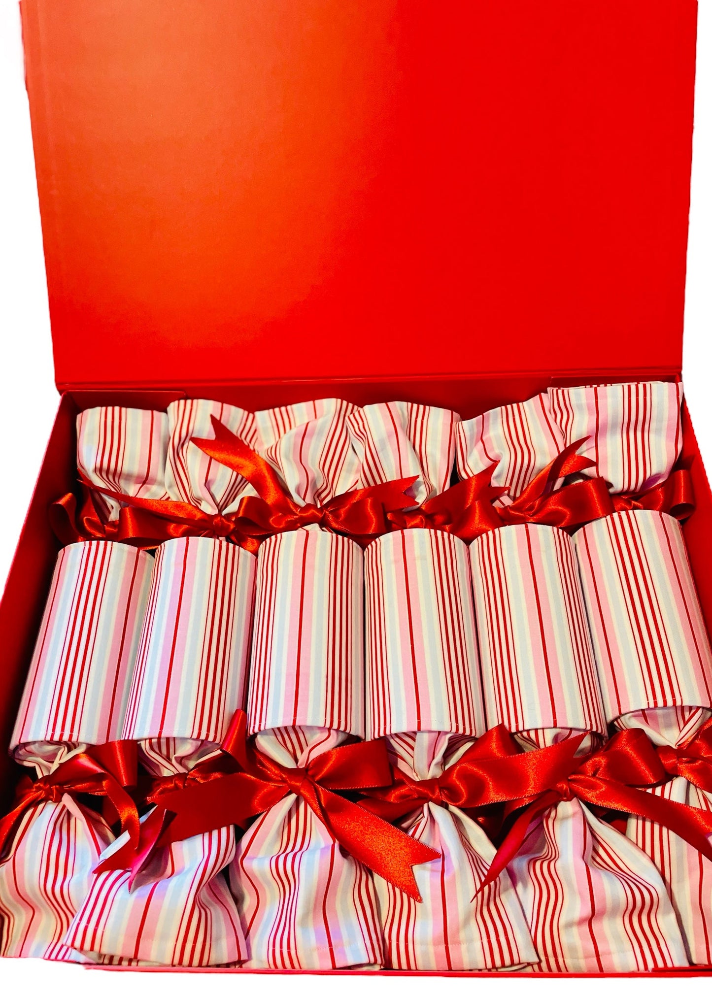 A set CANDY POP luxury Christmas crackers with white, red, and pink stripes, tied with red ribbons, displayed on a wooden surface with green foliage in the background.