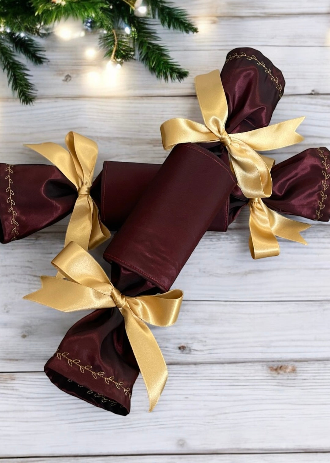 “Pair of Bordeaux satin reusable Christmas crackers with gold satin bows and embroidered edges on festive table setting”