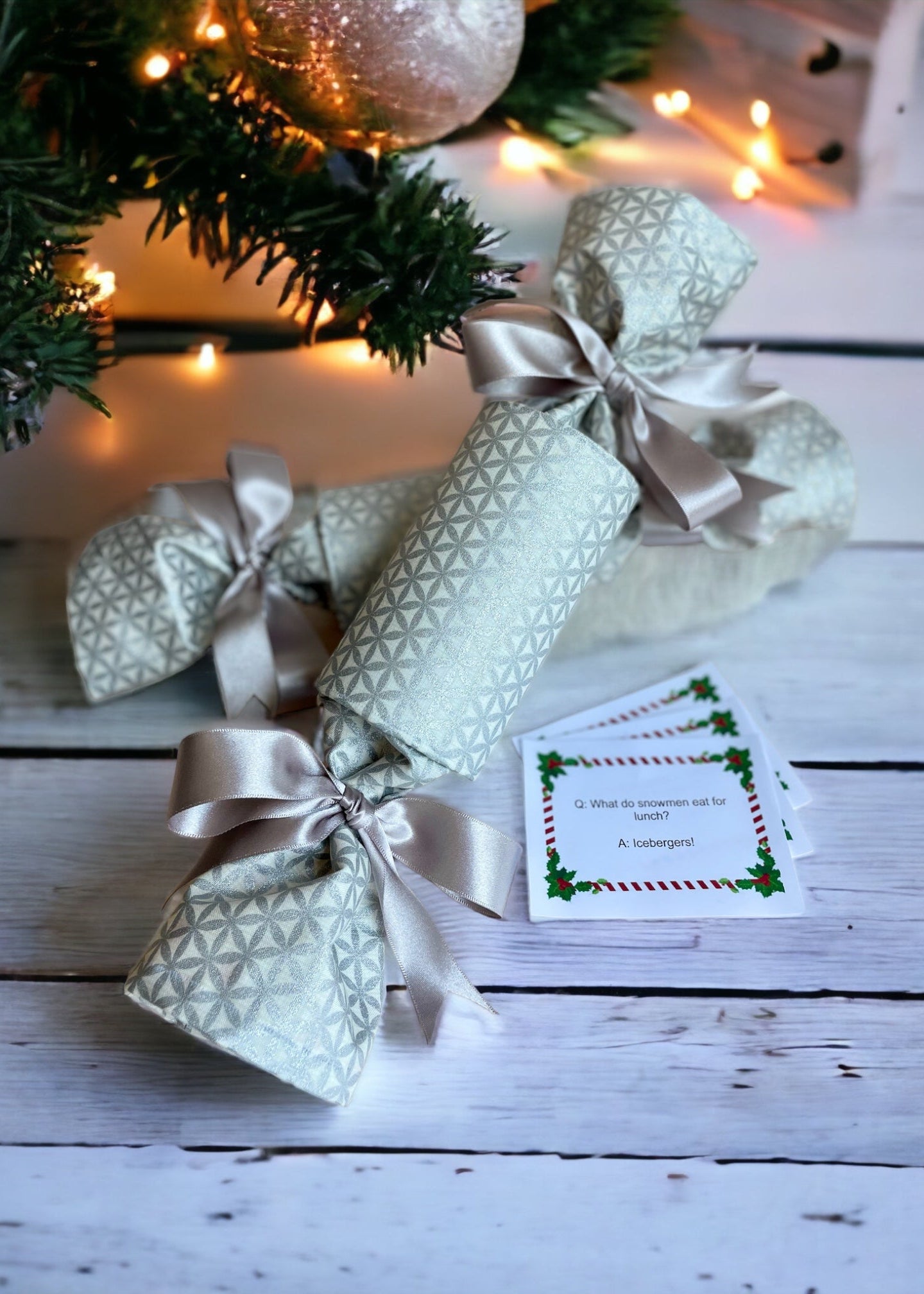 A set of reusable luxury Christmas crackers with a silver shimmer abstract pattern, tied with silver ribbons, displayed on a white wooden surface with Christmas decorations in the background.