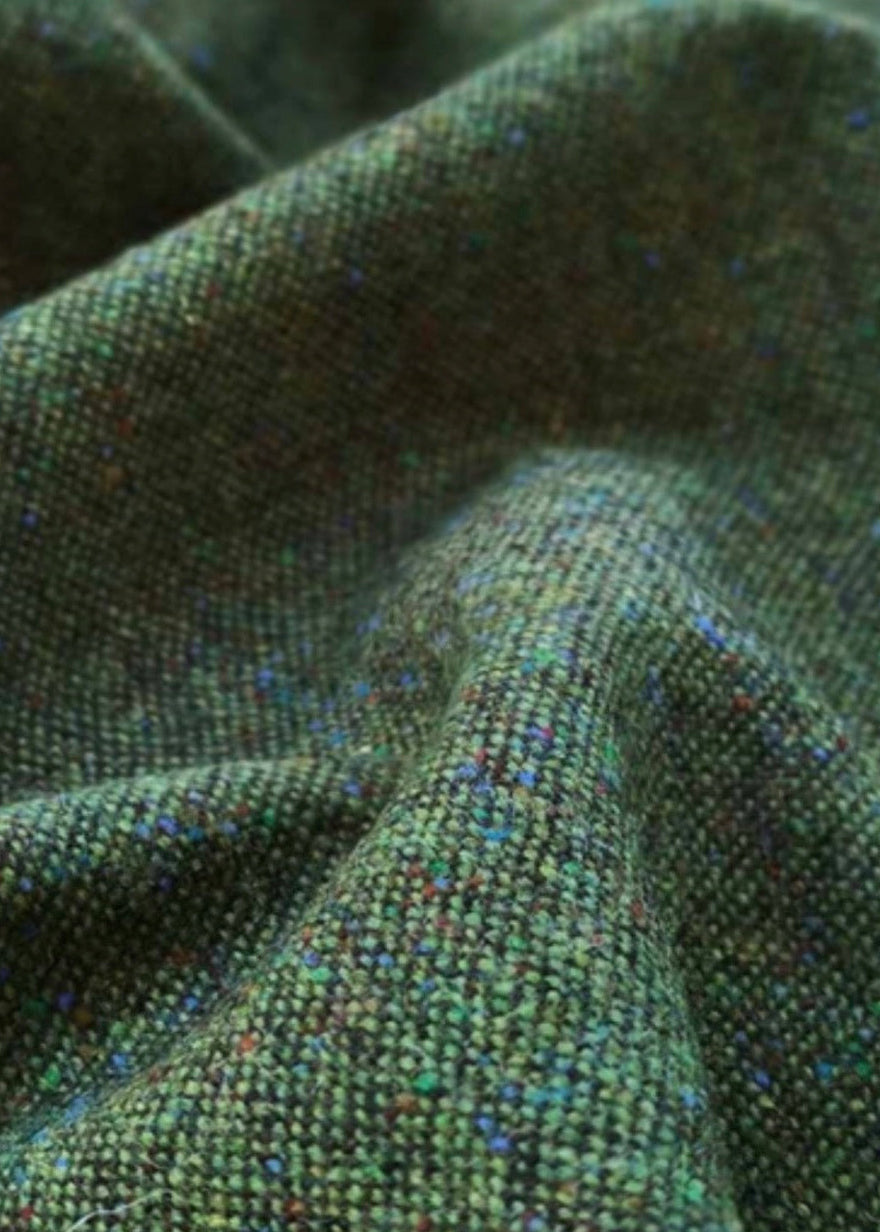 Green Donegal tweed fabric used to make reusable Christmas crackers, showing a coarse woven texture with dark and light green threads interlaced with small salt-and-pepper flecks of blue, red, teal, cream, and black.